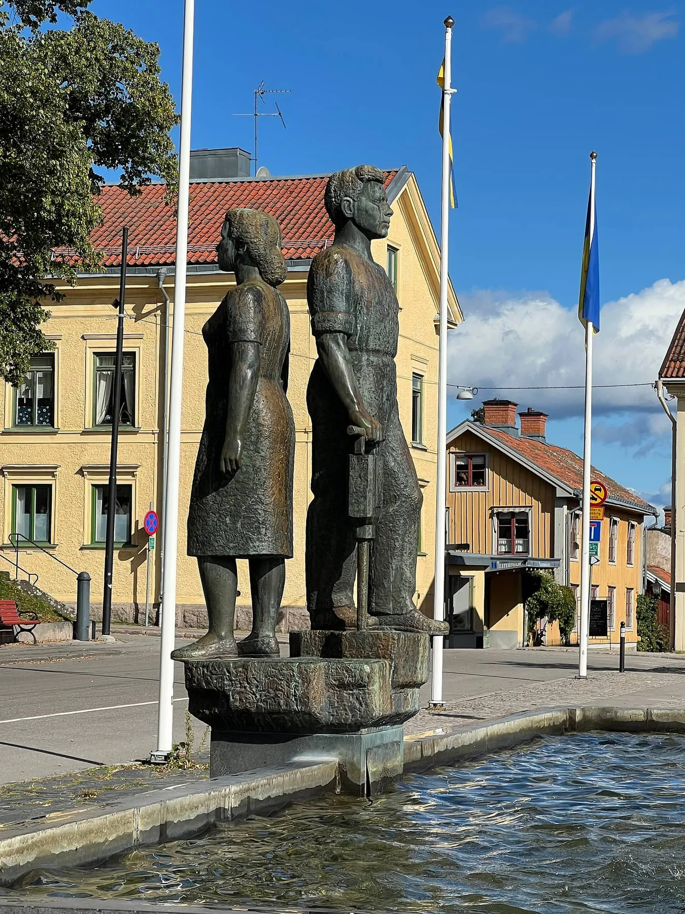Bronsskulptur av två stående figurer i en fontän på Nora torg, med gula och vita hus och flaggstänger bakom