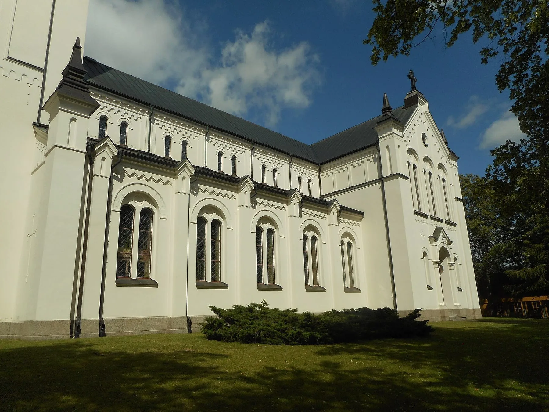 Vitputsad kyrka med höga rundbågefönster och svart tak mot blå himmel, sedd snett från sidan över gräsmattan