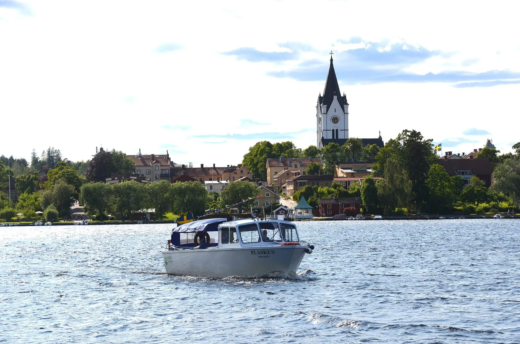 Liten vit passagerarbåt på Norasjön, med Noras bebyggelse och det höga vita kyrktornet på stranden bakom