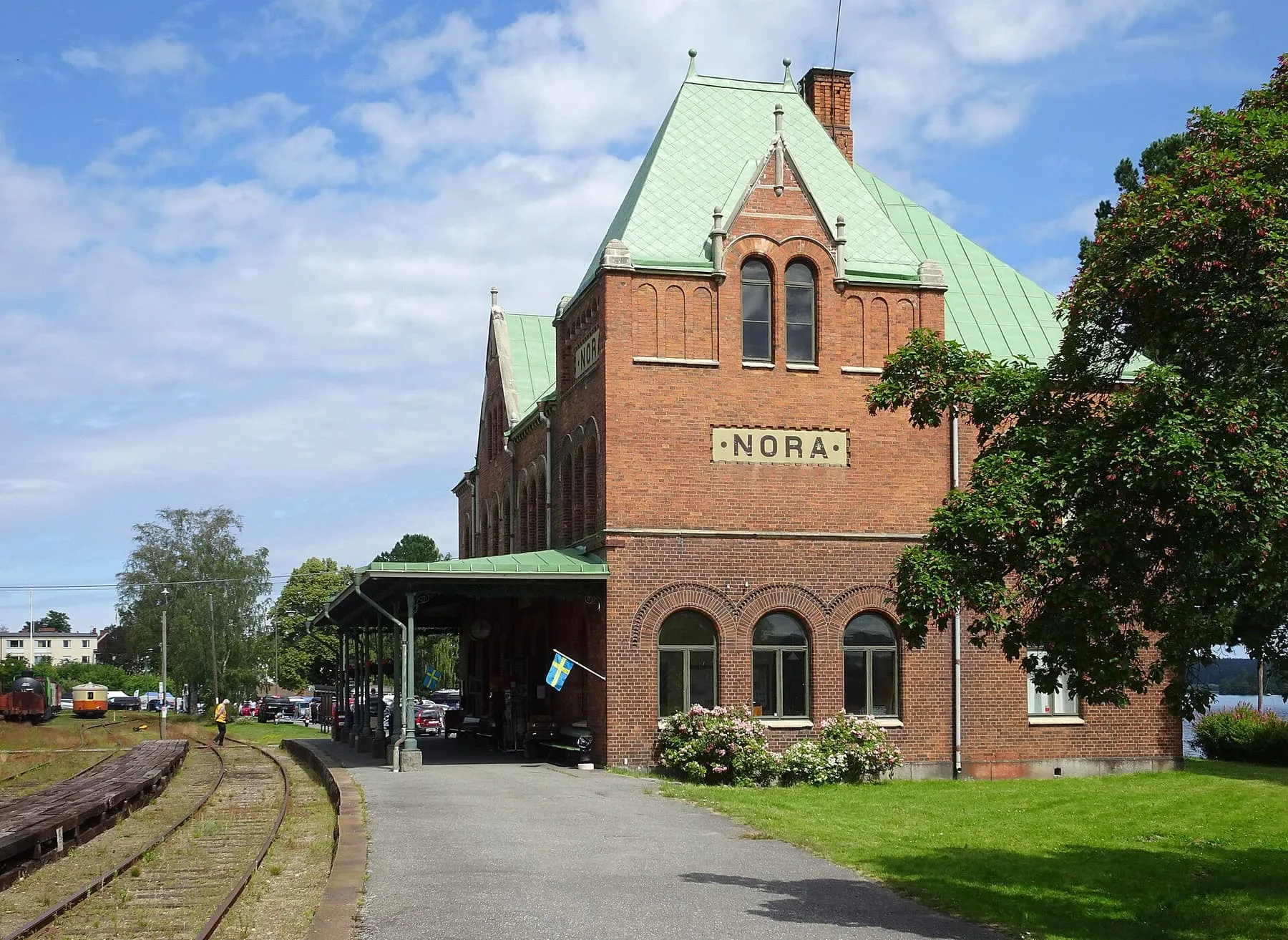 Stationshus i rött tegel med grönt plåttak och skylten Nora, med spår och perrong i förgrunden