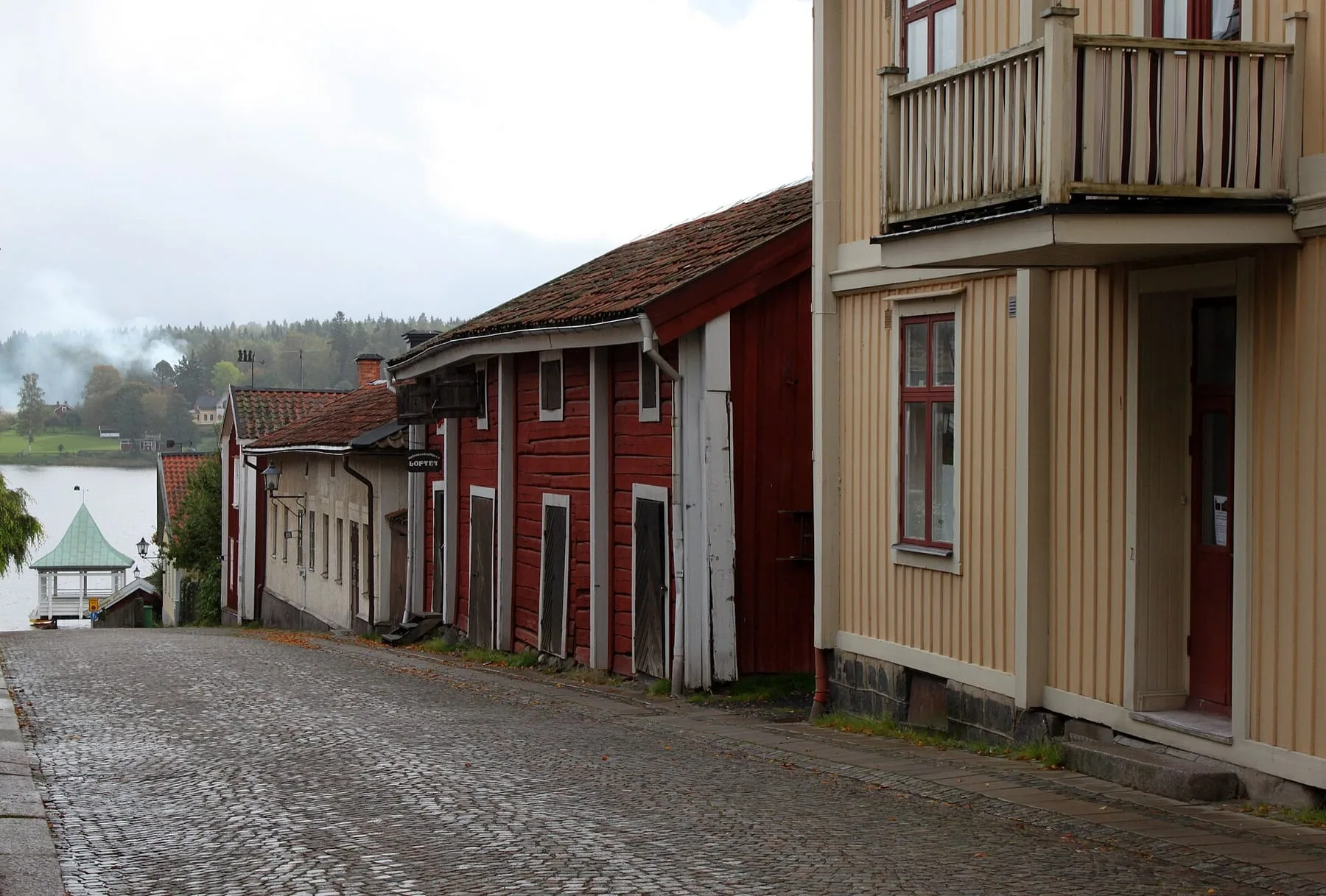 Smal kullerstensgata mellan röda och gula trähus i Nora, med en glimt av Norasjön i slutet av gatan