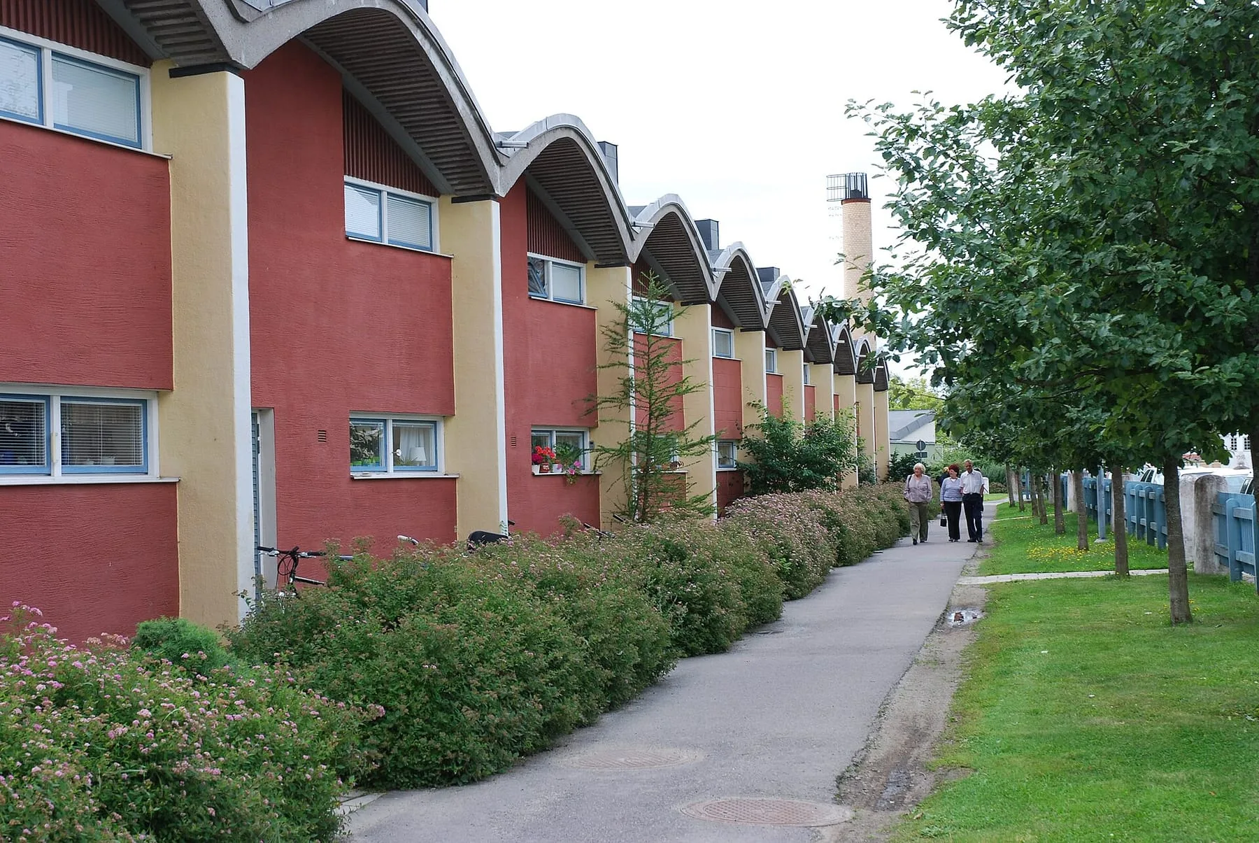 Lång radhuslänga i rött och gult med böjda tak längs en gångväg kantad av buskar