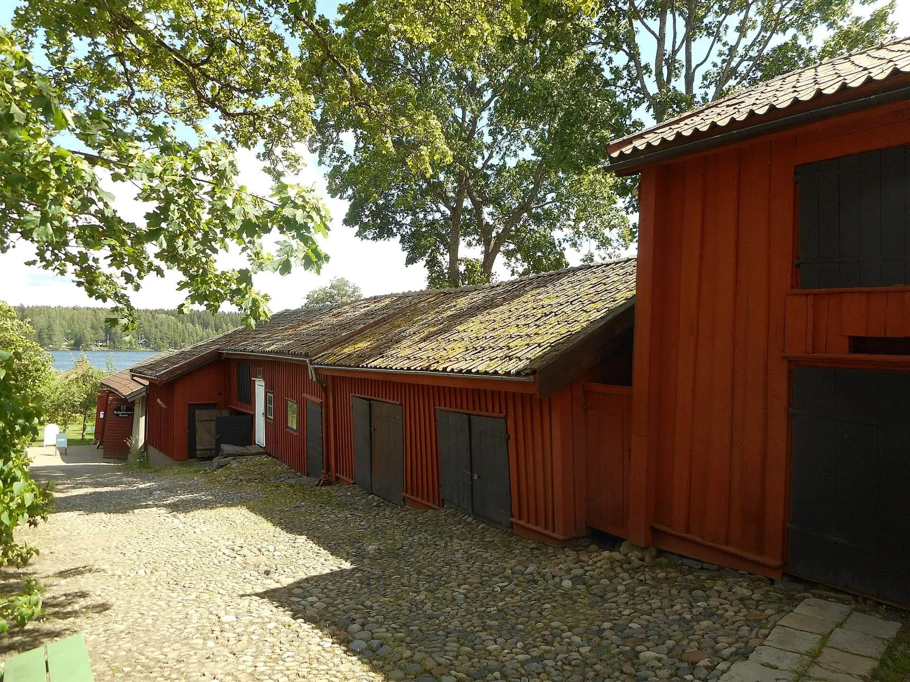 Kullerstensgård med låga faluröda uthus och ett skymt av Norasjön mellan träden.