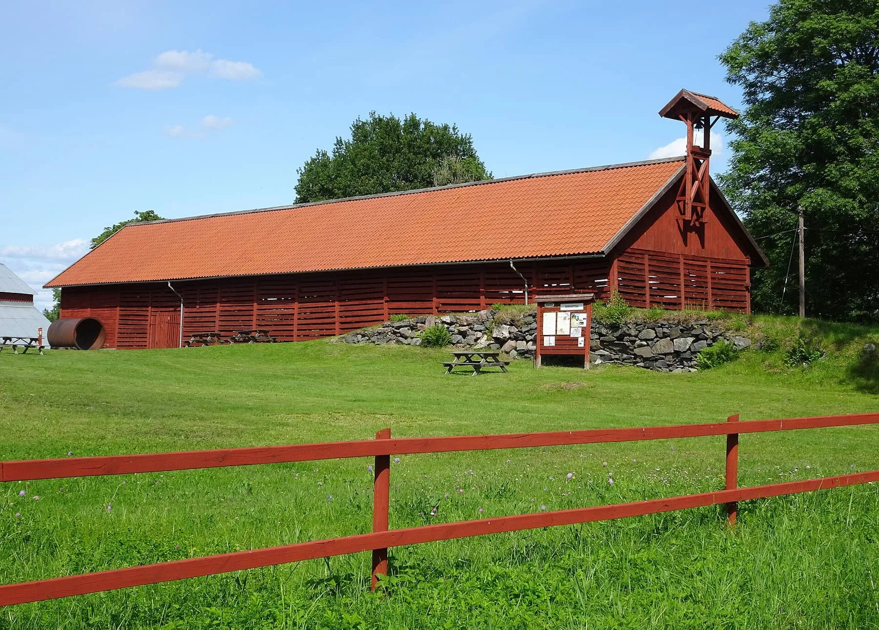 Långt rött kolhus med tegeltak och en liten klockstapel bakom ett rött staket på en äng